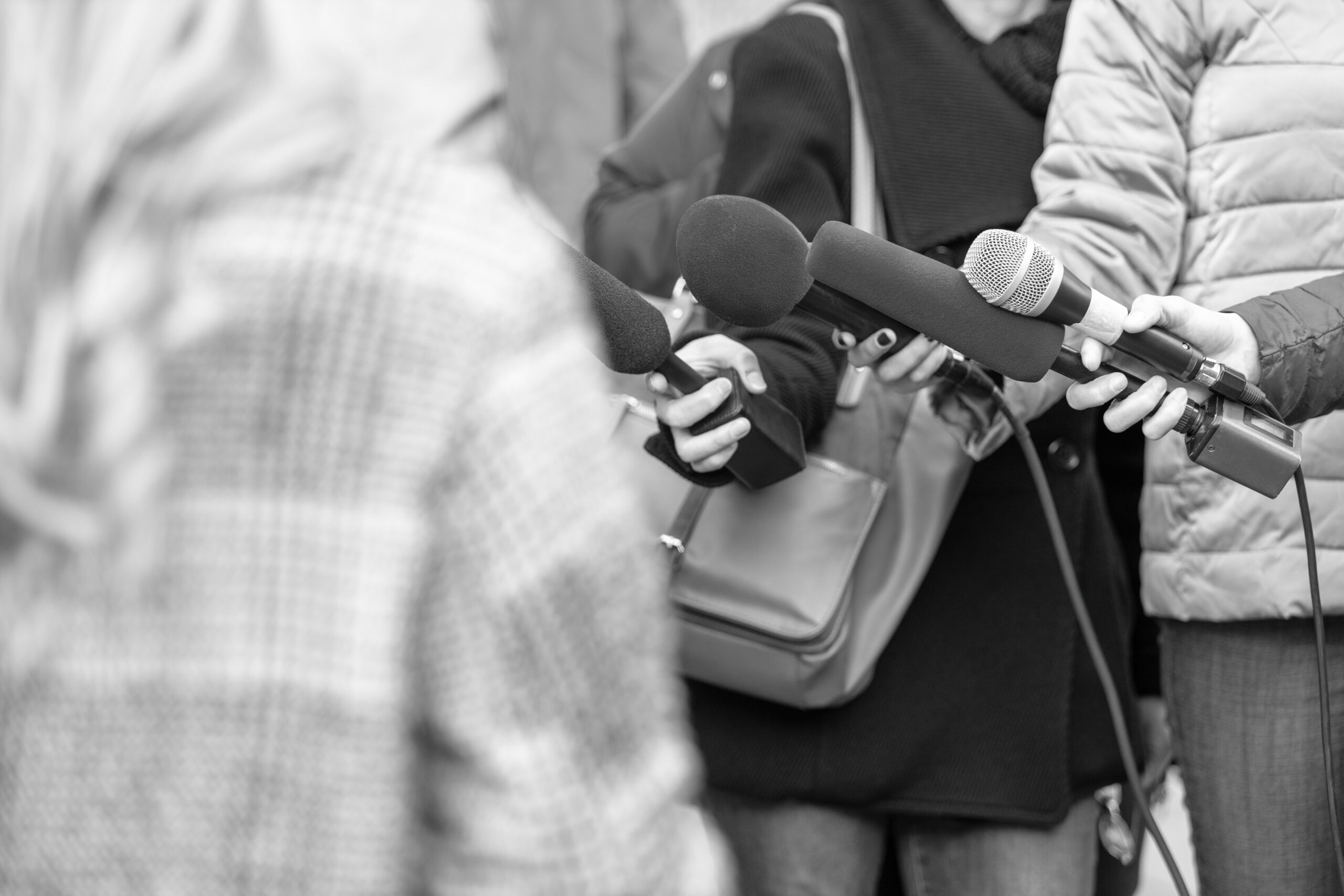 <p>Journalists holding microphones interviewing spokesperson outdoors, media scrum concept, breaking news, press conference, public relations, journalism, communication, reportage, selective focus, monochrome image</p>
